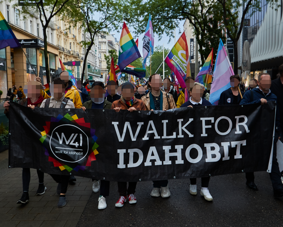 Eine Reihe von Personen hält ein Banner auf welchem "Walk for IDAHOBIT" zweimal steht. Hinter diesen sind weitere Menschen und einige Regenbogen sowie transfarbene Flaggen.